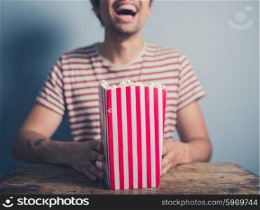 A happy laughing young man is sitting at a table with a box of popcorn