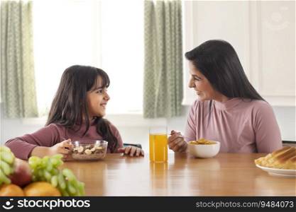 A HAPPY MOTHER AND DAUGHTER LOOKING AT EACH OTHER WHILE EATING BREAKFAST