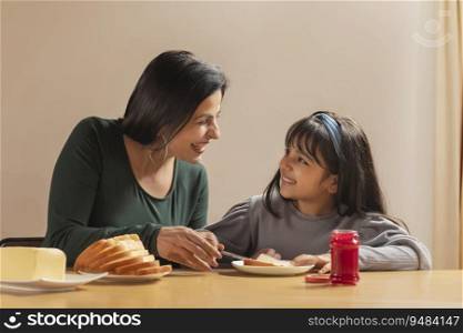 A HAPPY MOTHER LOOKING AT DAUGHTER WHILE SPREADING JAM ON A SLICE OF BREAD