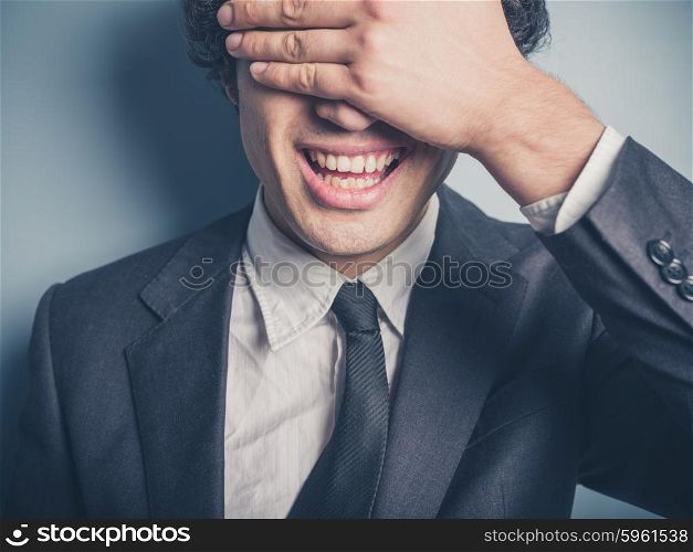 A happy young businessman is covering his face and eyes with his hand