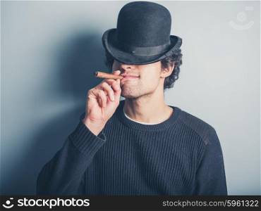 A happy young man wearing a black sweater and a bowler hat is smoking a cigar