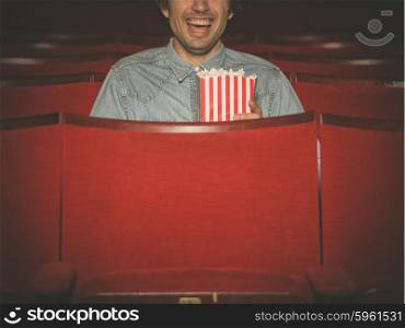 A happy young man with a bucket of popcorn is laughing and watching a movie at the cinema
