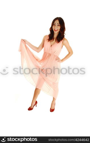 A happy young woman in a light pink dress, lifting the dress up, with redhigh heels and long brunette hair, standing for white background.