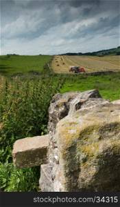 A hay baler at work in a field in the countryside, with a rock wall in the foreground