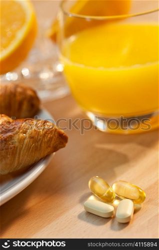 A healthy continental breakfast of croissant pastries, orange juice, oranges and in focus in the foreground are vitamin tablets and cod liver oil capsules, Illuminated with golden early morning sunshine.