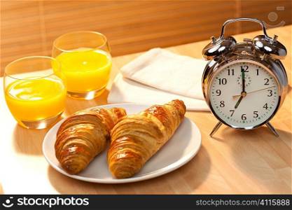 A healthy continental breakfast of croissant pastries, orange juice, oranges Illuminated with golden early morning sunshine and accompanied by a classic alarm clock set at 7am