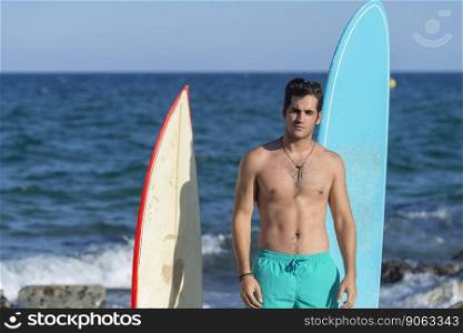 A Hispanic surfer guy standing on the beach in a swimsuit looking at the camera next to two surfboards.