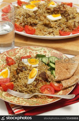 A homemade traditional Indian beef biryani, served with hard-boiled eggs, tomato and cucumber, with flat bread and a glass of water, and the serving dish in the background