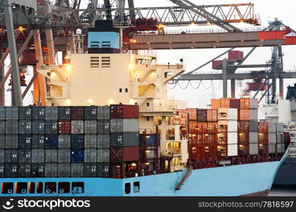 A huge container ship being unloaded at dawn in an industrial harbor by overhead cranes