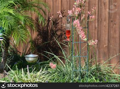 A hummingbird feeder filled with red syrup in the landscaped flower bed in a back yard.