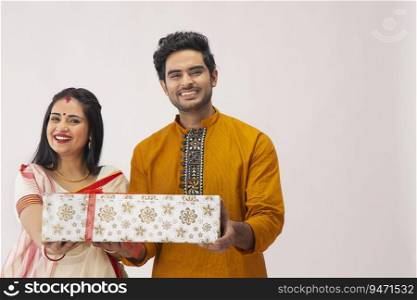 A HUSBAND AND WIFE LOOKING AT CAMERA WHILE HOLDING A GIFT