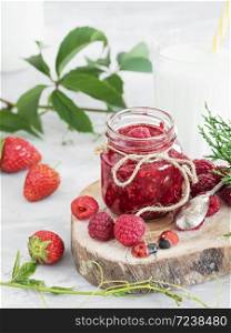 A jar of jam is located on a wooden plate, with fresh berries lying around. In the background is a glass of milk. The table is decorated with ivy and thuja branches. Gray background. Close-up.. A jar of jam is located on a wooden plate, with fresh berries lying around. In the background is a glass of milk. The table is decorated with ivy and thuja branches. Breakfast Close-up.