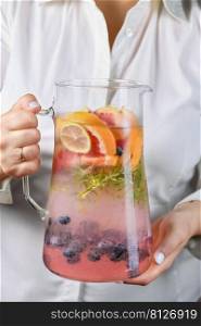 A jug with a refreshing organic soft drink with lime, blueberry, grapefruit and rosemary in female hands close-up