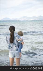 A lady holding a toddler standing in front of the sea with mountains in the far background