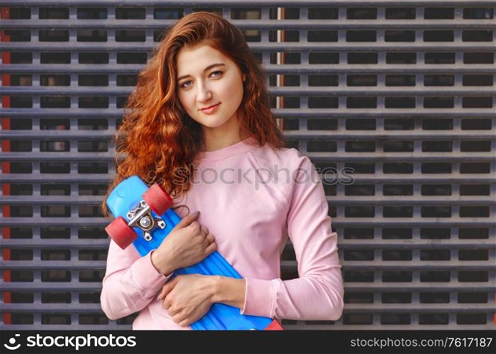 A Lady with long curly brown hair holds a blue skateboard in front of the chest. She stands with blank expression on her face in front of an iron grating in a skateboard park. Vintage color image.. Lady with long curly red hair holds a blue skateboard in front of the chest. She stands with blank expression on her face in front of an iron grating in a skateboard park. Vintage color.
