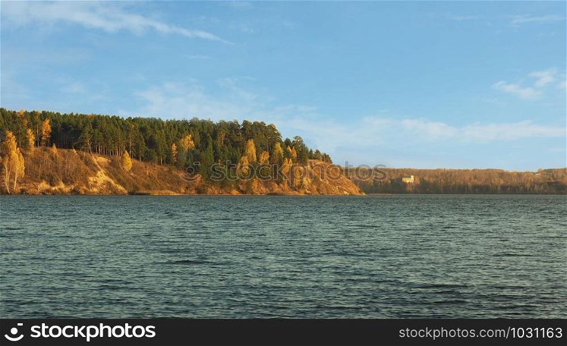 A lake with shallow waves on the surface and an autumn forest on the opposite bank. Blue sky with clouds.