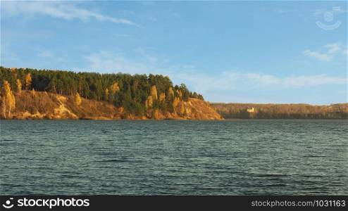 A lake with shallow waves on the surface and an autumn forest on the opposite bank. Blue sky with clouds.