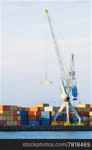 A large cargo crane stands at port in front of stacks of containers. There is no one viewable in the image. Vertically framed shot.