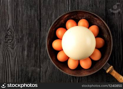 A large ostrich egg surrounded by chicken eggs in an old cast-iron pan that stands on an old black wooden surface, low key image, top view, copy space.. Large ostrich egg surrounded by chicken eggs in an old cast-iron pan, close-up.