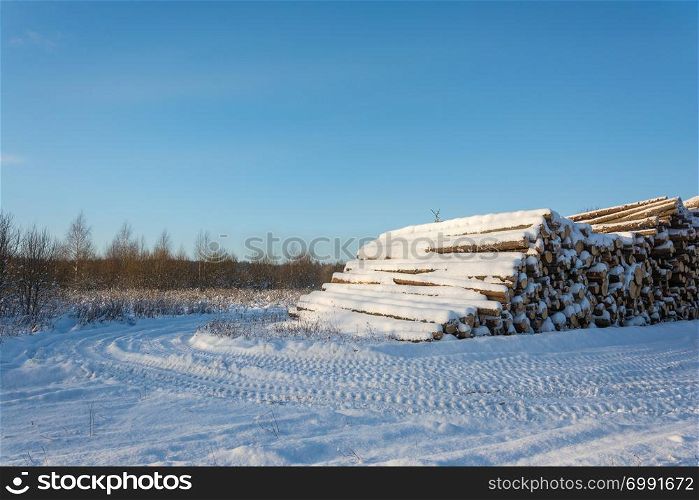 A large pile of snow covered logs on the edge of the road on a clear winter day.