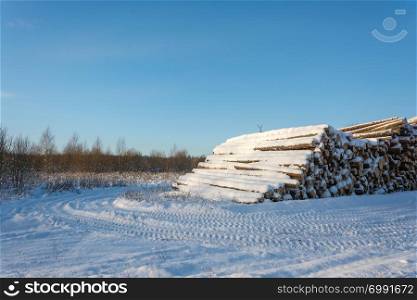 A large pile of snow covered logs on the edge of the road on a clear winter day.