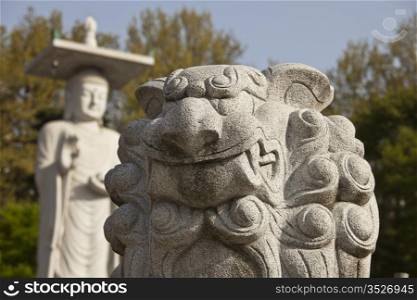 A lion carved out of white marble stands in front of a large, imposing figure of Buddha at an outdoors worship area in the Bongeunsa temple in Seoul, South Korea.