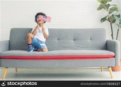 A little african black boy is waving american flag while sitting on sofa in living room at home. Diversity and Black lives Matter Concept.