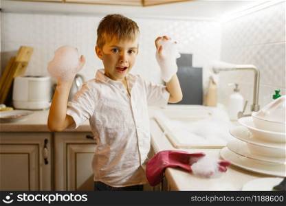 A little boy in gloves washing dishes on the kitchen. Baby doing housework at home. Young mom&rsquo;s helper cleans the house. Little boy in gloves washing dishes on the kitchen