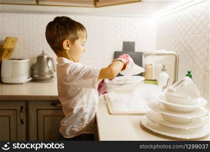A little boy in gloves washing dishes on the kitchen. Baby doing housework at home. Young mom&rsquo;s helper clean the house. Little boy in gloves washing dishes on the kitchen