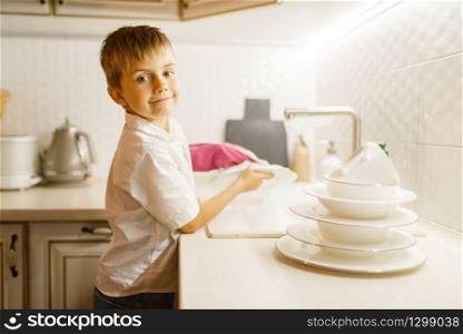 A little boy in gloves washing dishes on the kitchen. Baby doing housework at home. Young mom&rsquo;s helper clean the house