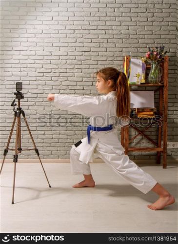 A little girl in a white kimono with a blue belt takes karate lessons via the Internet while at home on self-isolation during quarantine. karate girl internet