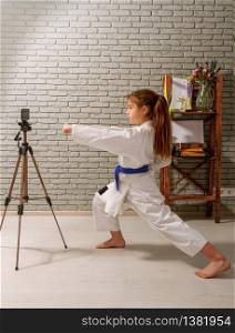 A little girl in a white kimono with a blue belt takes karate lessons via the Internet while at home on self-isolation during quarantine. karate girl internet
