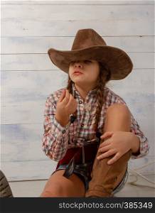 A little girl in a wide-brimmed cowboy hat wearing a traditional dress and high boots with a lasso is eating a straw on a light wooden background