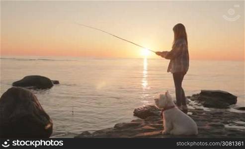 A little girl is fishing with a fishing rod on the sea coast. A small dog is sitting next to it. Sunset at the sea. Slow motion