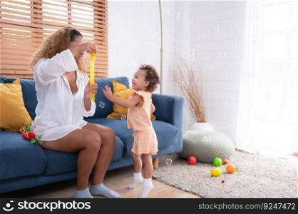 A little girl runs and plays with a small soap bubble that his mother blows for him to play with. In the living room of the house