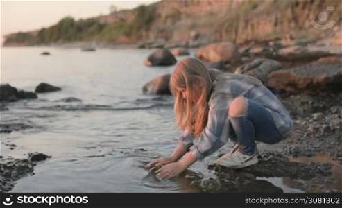 A little girl washes her hands and face with sea water by the sea coast. Sunset at the sea.