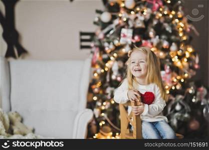 A little girl with a red heart on the clothes sitting in front of the tree.. The girl with big red heart on the clothes near the Christmas tree 7302.