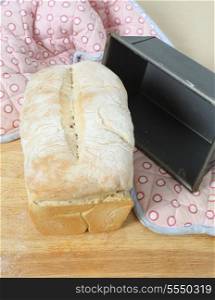 A loaf of English split-tin bread, fresh from the oven, on a wooden board with the baking tin and oven gloves, vertical orientation.