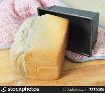 A loaf of English split-tin bread, fresh from the oven, on a wooden board with the baking tin and oven gloves.