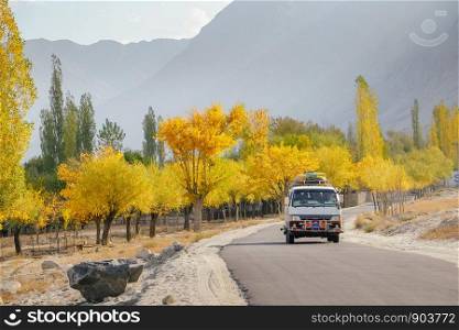 A local bus running on paved road with luggage bags on the roof with morning sunlight. Landscape view of colorful trees in autumn against mountain range in Skardu. Gilgit Baltistan, Pakistan.