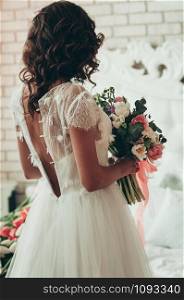 A long haired brunette bride in unbuttoned white dress with wedding bouquet, a view from the back
