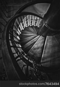 A look down to an old spiral staircase. Wooden circular stairway with ornate metallic railing, black and white vertical shot, interior architecture details of stair steps inside an ancient building.