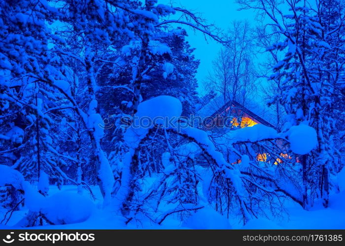 A lot of snow in the evening winter forest. Lonely wooden cottage glows in the thicket. Evening in a Snowy Forest and a Lighted Cottage