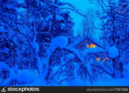 A lot of snow in the evening winter forest. Lonely wooden cottage glows in the thicket. Evening in a Snowy Forest and a Lighted Cottage
