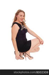 A lovely blond woman in a black dress and high heels crouching on the floor smiling into the camera, isolated for white background.