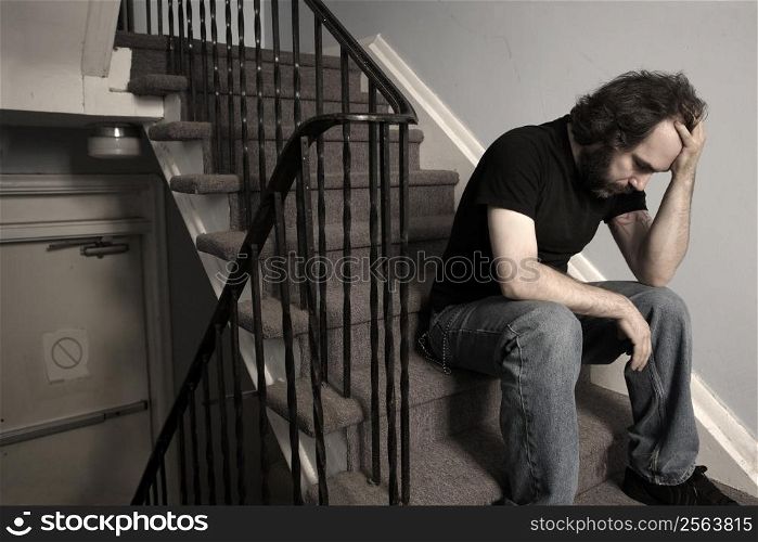 A male adult with overwhelming depression sitting in the stairwell of his apartment building. Desaturated.