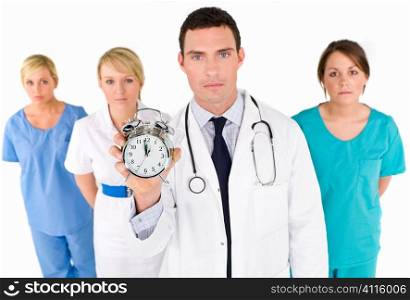 A male doctor backed by his medical team holding out an alarm clock ticking ever closer to 12 o&acute;clock. The focus is on the clock face.