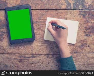 A male hand is writing in a notepad there is a tablet computer on the table as well with a green screen