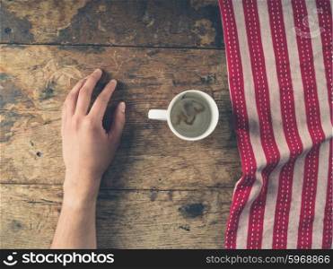 A male hand resting on a wooden table with a red and white tea towel and an empty cup