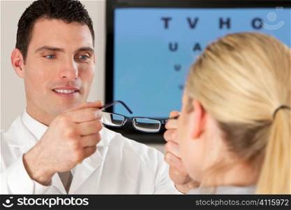 A male optician fitting glasses onto a blond female patient with an electronic eyechart out of focus behind him.